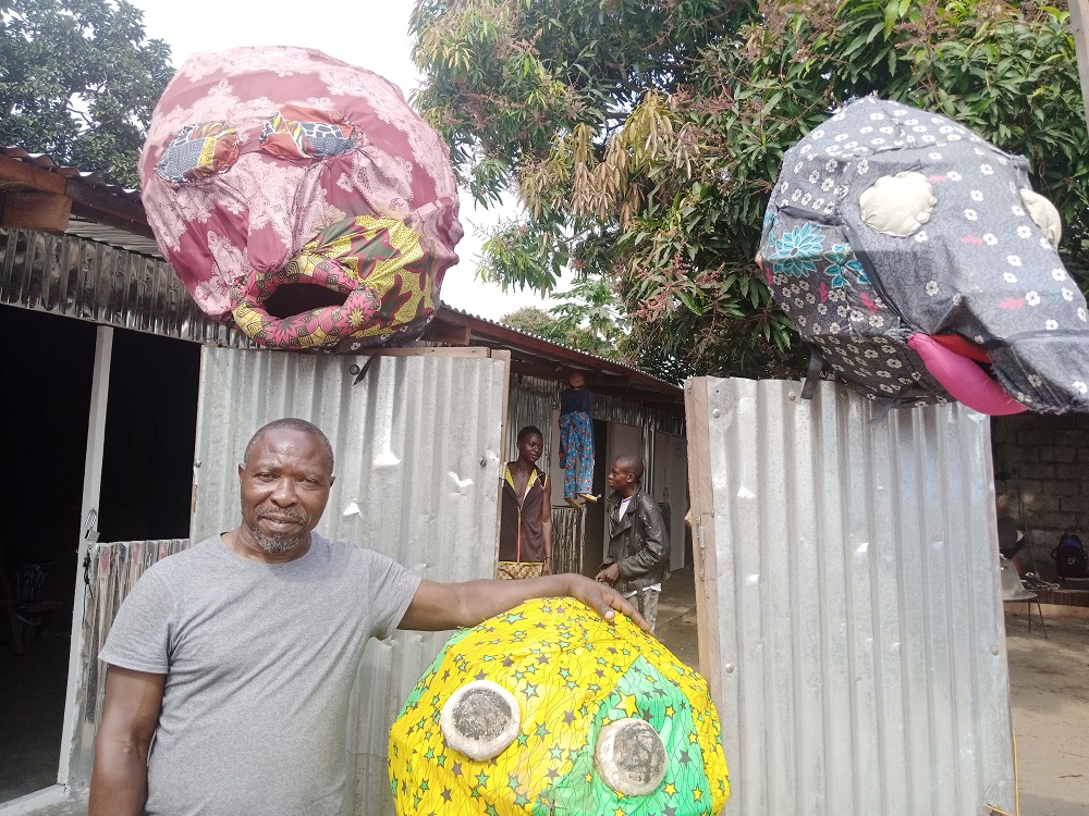 Hubert Mahela posant devant des têtes de marionnettes géantes de l’Espace Masolo (Adiac)